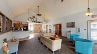 This single-floor community clubhouse is designed to offer a welcoming and versatile gathering space. The main lounge area immediately impresses with dramatic vaulted ceilings and dual ceiling fans, creating an airy, open atmosphere. The space is furnished with neutral-toned sectional seating and vibrant turquoise accent chairs arranged around a central coffee table, offering comfortable areas for both casual conversation and relaxation. A stone-faced fireplace anchors one wall, accompanied by a wall-mounted flat-screen television and flanked by large windows that invite an abundance of natural light and views of the surrounding outdoor area. Bar-height tables and stools nearby provide additional seating options. A reception or service counter with a dark granite surface and hardwood flooring marks the transition toward the kitchenette, which features wood cabinetry, a coffee station, and standard appliances. A pass-through window connects this area to an adjacent room. French doors with warm wood trim open to additional spaces, enhancing the flow and connectivity throughout. A charming dining nook, accessed through one of these doors, features an exposed brick accent wall with recessed lighting above a round wooden dining table, lending a warm, intimate character. Throughout the space, patterned carpet in the lounge areas and hardwood flooring near the service counter complement the overall inviting ambiance of this well-appointed facility.