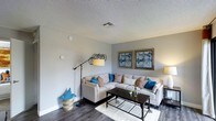 This single-floor space features a thoughtfully designed living room that serves as its centerpiece. The room is finished with durable wood-look flooring in a warm gray tone, providing a contemporary foundation that complements the neutral color palette of the walls. A textured popcorn ceiling adds architectural character overhead, while a centrally located HVAC vent ensures climate comfort throughout.

The living area is anchored by a large L-shaped sectional sofa in a light beige upholstery, dressed with an eclectic mix of decorative pillows in shades of blue, orange, and white. A dark-framed rectangular coffee table sits atop a plush white area rug, grounding the seating arrangement. A pair of framed blue-toned abstract artworks mounted on the far wall introduce a bold visual focal point, echoing the blue accent tones present in the soft furnishings.

Lighting is provided by a tall adjustable arc floor lamp and a warm table lamp, creating a layered and inviting ambiance. A decorative woven basket and sculptural wall accent add finishing touches, giving the space a curated, lived-in feel that balances comfort with modern style.