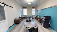 This single-floor space presents a well-appointed lounge or common area designed for comfort and functionality. The interior features a neutral color palette of soft beige and warm white walls, accented by a bold teal feature wall that adds a vibrant focal point to the room. Crown molding along the ceiling lends a polished, refined touch to the overall aesthetic.

The space is furnished with multiple seating arrangements, including plush brown upholstered armchairs and small round tables with black chairs, encouraging both individual relaxation and small group interaction. Along the walls, a set of mint-green cushioned chairs provides additional casual seating near the windows. Natural light flows generously through multiple windows dressed with warm orange roller shades, creating a bright and inviting atmosphere.

Light wood-toned flooring with subtle striping runs throughout, complementing the contemporary furnishings and adding warmth to the space. A teal accent strip along the base of one wall echoes the feature wall and ties the color scheme together cohesively. Office equipment and storage units are neatly positioned along one side, suggesting a dual-purpose environment suited for both relaxation and productivity. Decorative elements, including a tall vase with dried florals, add a finishing touch of elegance to this functional yet welcoming space.