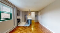 Located at 12 Marshall Street in Irvington, NJ, this efficiently designed unit makes excellent use of its single-floor layout. The combined kitchen and living area forms the heart of the space, where warm parquet flooring flows throughout, lending a classic yet timeless character to the interior. The kitchen is well-appointed with stainless steel appliances, including a gas range, over-the-range microwave, and refrigerator. Gray wood-tone cabinetry extends to the ceiling, providing ample storage, while a sleek white countertop breakfast bar serves as a natural divider between the kitchen and living area. Amber pendant lights hang above the bar, adding a warm, decorative touch. Natural light enters the living area through a window fitted with blinds, and a baseboard heating unit runs along the wall beneath it. The neutral gray walls throughout create a clean, modern backdrop. The bathroom offers a refined finish with marble-style wall surround encasing the bathtub and shower combination, complemented by a sliding glass enclosure and a clean white basin. A hallway connects the main living space to the entry, which features a utility panel and a solid white entry door, completing this practical and attractively updated unit.