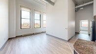 Located in Pittsburgh's South Side neighborhood, this two-bedroom, one-bathroom unit offers an open, industrial-inspired layout across a single floor. The main living area immediately impresses with its soaring ceilings, exposed round ductwork, and large multi-pane windows that flood the space with natural light while framing views of the surrounding urban landscape. Light-toned hardwood-style flooring runs throughout the living area, complementing the bright, neutral wall palette. The kitchen is integrated into the open-plan living space and features dark cabinetry, a granite-topped island with a rich red finish, stainless steel appliances, and pendant lighting overhead, adding warmth and contrast to the airy surroundings. Both bedrooms are carpeted and well-sized, with the primary bedroom offering notable square footage. Each bedroom benefits from windows with roller shades and baseboard heating units, ensuring comfort year-round. The shared full bathroom features wood-look flooring, a full tub-shower combination, and an adjacent closet for added storage. A hallway connects the private areas of the unit to the main living space, providing a clear and functional layout throughout.