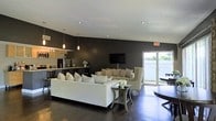 This single-floor space showcases a striking open-concept design that seamlessly connects the living, dining, and kitchen areas into one cohesive, modern environment. The interior is defined by dramatic dark walls that create a bold contrast against the light-toned furnishings, including a plush white sectional sofa, neutral-toned accent chairs, and decorative throw pillows that add warmth and texture throughout. Recessed lighting and sleek pendant lights suspended from the textured white ceiling provide layered illumination, enhancing the space's contemporary ambiance. Rich, dark hardwood flooring runs throughout, anchoring the sophisticated color palette. The kitchen area features a breakfast bar with bar seating, pendant lighting, and stainless steel appliances, making it both functional and visually appealing. A wall-mounted flat-screen television serves as a focal point in the living area. Large sliding glass doors flood the space with natural light and offer a direct view to an outdoor area, creating an effortless indoor-outdoor connection. The overall atmosphere is polished and stylish, with a carefully curated balance of dark and light tones that lend the space a refined, upscale character.
