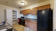 This single-floor residence offers a thoughtfully arranged layout spread across a generously sized footprint. The living area is notably open and expansive, featuring neutral-toned carpet, a ceiling fan with light fixture, and a gray accent wall that adds a touch of visual contrast. Large windows with vertical blinds allow ample natural light to fill the space, while a sliding glass door provides access to an outdoor area. A bifold-door closet adds convenient built-in storage directly within the living space.

The kitchen is functional and well-appointed, featuring warm-toned wood cabinetry extending to the ceiling for substantial storage, dark granite-style countertops, and a full suite of black appliances including a gas range, dishwasher, and refrigerator. A double basin sink and a separate walk-in pantry closet with open shelving further enhance the kitchen's practicality. Tile flooring completes the kitchen area.

The bedrooms are carpeted and receive natural light through windows with vertical blinds. Each room offers a clean, neutral palette suitable for versatile use.

The bathroom features full-height beige tile surrounding a soaking tub and shower combination, complemented by a wood-toned vanity with a white countertop and a large mirror above, creating a clean, cohesive aesthetic throughout.
