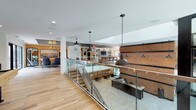 This thoughtfully designed amenity space combines industrial and contemporary elements across a dynamic, open layout with distinct functional zones. The main level is anchored by a sunken lounge area visible from an upper walkway, where sleek glass panel railings with stainless steel frames provide both safety and unobstructed sightlines. Exposed brick accent walls run throughout, adding warmth and texture that contrast with clean white ceilings and wood-look tile flooring.

The lounge area features a dark leather sofa, bold orange accent chairs, a large square coffee table, and curated black-and-white photographic artwork arranged in a gallery-style wall display. A mid-century-inspired chandelier and recessed lighting illuminate the space comfortably. Adjacent to the lounge, a high-top dining area offers round pub tables with yellow-orange metal barstools positioned alongside a wide horizontal window, providing natural light and views of the surrounding greenery.

The kitchenette is outfitted with flat-panel light wood cabinetry, stone countertops, a stainless steel sink, built-in appliances, and a sea-glass blue subway tile backsplash with a wall-mounted television above. Industrial-style pendant lights hang throughout the upper level, reinforcing the cohesive aesthetic. Overall, the space delivers a welcoming, social atmosphere suited for gathering and everyday use.