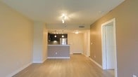 This single-floor unit offers a clean, modern design with a thoughtful open-concept layout that seamlessly connects the main living area to the kitchen. Light hardwood flooring runs throughout, lending warmth and continuity to the space, while neutral-toned walls create a bright and welcoming ambiance.

The kitchen is a standout feature, showcasing dark cabinetry that contrasts elegantly with a raised breakfast bar peninsula finished in a complementary tone. Stainless steel appliances, including a refrigerator and microwave, add a contemporary touch, while pendant lighting suspended above the peninsula provides both functional illumination and a stylish accent. A tile backsplash completes the kitchen's polished look.

The open living area is generously proportioned, bathed in light from ceiling-mounted fixtures and benefiting from the open sightlines the layout affords. A dedicated entryway with a clean white door provides a clear sense of arrival and separation from the main living space.

Throughout the unit, clean white baseboards and trim offer a crisp finish, and ceiling vents ensure efficient climate control. The overall design strikes a balance between modern aesthetics and practical livability, making it a well-appointed and functional space.