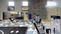 Located in Silverdale, WA, this single-floor fitness center offers a thoughtfully designed workout environment that balances open space with defined functional zones. The facility is characterized by soaring vaulted ceilings equipped with skylights and recessed lighting, flooding the interior with natural and artificial light throughout the day. High clerestory windows further enhance brightness while maintaining an energetic atmosphere.

The main exercise floor is covered in a bold geometric tile pattern in blue, gray, and white tones, creating a visually dynamic foundation. A dedicated functional training zone features a dark sport-turf mat printed with track-style lane markings, anchored by a central multi-use cable and strength rig. This area accommodates battle ropes, medicine balls, and suspension training accessories.

Surrounding the functional zone, a comprehensive selection of Life Fitness selectorized strength machines is arranged to allow easy circulation. A cardio section houses treadmills and steppers positioned near wall-mounted flat-screen televisions. Full-length mirrors expand the sense of space and aid in form monitoring. A glass-partitioned area adjacent to the main floor provides visual connectivity while defining a separate training enclosure.

An exposed brick accent wall with a large window frames views of surrounding greenery, adding warmth and texture to the otherwise contemporary interior. A glass-railed staircase connects to an upper level, completing this well-equipped facility.