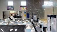 Located in Silverdale, WA, this single-floor fitness center offers a thoughtfully designed workout environment that balances open space with defined functional zones. The facility is characterized by soaring vaulted ceilings equipped with skylights and recessed lighting, flooding the interior with natural and artificial light throughout the day. High clerestory windows further enhance brightness while maintaining an energetic atmosphere.

The main exercise floor is covered in a bold geometric tile pattern in blue, gray, and white tones, creating a visually dynamic foundation. A dedicated functional training zone features a dark sport-turf mat printed with track-style lane markings, anchored by a central multi-use cable and strength rig. This area accommodates battle ropes, medicine balls, and suspension training accessories.

Surrounding the functional zone, a comprehensive selection of Life Fitness selectorized strength machines is arranged to allow easy circulation. A cardio section houses treadmills and steppers positioned near wall-mounted flat-screen televisions. Full-length mirrors expand the sense of space and aid in form monitoring. A glass-partitioned area adjacent to the main floor provides visual connectivity while defining a separate training enclosure.

An exposed brick accent wall with a large window frames views of surrounding greenery, adding warmth and texture to the otherwise contemporary interior. A glass-railed staircase connects to an upper level, completing this well-equipped facility.