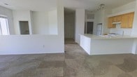 This single-floor space is designed with an open-concept layout that integrates the living area and kitchen into one cohesive, flowing environment. The interior is finished in crisp white walls that enhance the sense of brightness and airiness throughout. The living area features wall-to-wall neutral-toned carpeting that adds warmth underfoot and complements the clean aesthetic of the space. A low half-wall partition gently defines the transition between the living and kitchen areas while maintaining an open and connected feel. The kitchen is anchored by a white peninsula counter that provides ample workspace and a casual seating or serving area. A refrigerator is visible in the background, and track lighting above the kitchen area ensures the workspace is well-lit. A pass-through window or opening in the wall adds an architectural detail that reinforces the open and communicative design of the floor plan. Doorways lead to additional areas of the space, suggesting further rooms beyond the main living zone. The overall ambiance is clean, minimal, and move-in ready, offering a versatile blank canvas well-suited to a variety of interior styles and furnishing preferences.