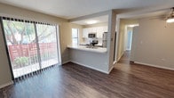 This single-story unit is thoughtfully laid out with an open-concept design connecting the living area and kitchen. Dark hardwood-style flooring runs throughout the main living spaces, creating a cohesive and modern aesthetic. The living area is generously sized and benefits from abundant natural light streaming through a large sliding glass door fitted with vertical blinds, which opens to a private patio with views of mature trees and a surrounding fence.

The kitchen is positioned at the heart of the unit behind a peninsula-style counter with a double-basin stainless steel sink and light-toned countertops. White upper cabinetry contrasts with dark lower cabinets, while a classic white subway tile backsplash adds texture. Stainless steel appliances, including a refrigerator, electric range, and over-the-range microwave, complete the space.

The bedrooms are carpeted and well-lit, each featuring windows with horizontal blinds. At least one bedroom includes a reach-in closet with built-in shelving. A clean, white bathroom offers a tub-shower combination, a single-sink vanity with a large mirror, and tile flooring. A dedicated laundry area houses a full-sized washer and dryer alongside a water heater, adding practical convenience to the layout.