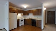 This single-floor space is designed with practicality and functionality in mind. The kitchen serves as a central highlight, featuring an abundance of dark wood cabinetry that offers generous storage both above and below the countertops. The L-shaped layout provides an efficient workflow, with ample counter space for meal preparation. Essential appliances are included, among them a white refrigerator, an electric range with an overhead ventilation hood, a dishwasher, and a double-basin sink with a standard faucet. The countertops are finished in a light tone that provides a clean contrast to the rich, warm hues of the surrounding cabinetry. The flooring throughout is a durable hardwood-style material in a warm brown finish, adding to the cohesive, grounded aesthetic of the space. The kitchen is lit by a flush-mount ceiling light, complemented by a textured ceiling that adds a subtle visual detail overhead. The overall ambiance of the space is straightforward and utilitarian, making it well-suited for everyday living. The neutral wall tones keep the environment feeling open and easy to personalize.