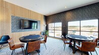 This dedicated game room is a thoughtfully designed social space that blends contemporary style with a comfortable, inviting atmosphere. The room accommodates two round poker-style tables, each surrounded by a mix of warm cognac leather chairs and modern molded side chairs, providing ample seating for group gaming sessions.

The interior design is defined by a striking contrast of materials: one wall features vertical wood slat paneling framing a large wall-mounted flat-screen television, while the remaining walls are adorned with a bold, dark botanical-patterned wallcovering that lends the space a sophisticated, moody character. Recessed lighting throughout the ceiling provides even illumination across the room.

Underfoot, a textured, patterned carpet adds visual interest and acoustic comfort. Multiple large windows span one wall, flooding the space with natural light and offering open views of the surrounding exterior landscape. Entry is through a set of elegant double doors with warm wood frames and glass panels, providing a clear sightline into the room.

Decorative touches include potted greenery and a sleek console table along the wall, adding warmth and life to the space. A wall-mounted HVAC unit ensures year-round climate comfort. Overall, this game room delivers a polished, modern environment ideal for social gatherings and recreational activities.