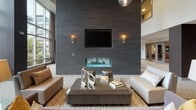 This elegantly designed single-floor amenity space exudes a contemporary, upscale ambiance from the moment you enter. The entryway welcomes visitors through sleek glass sliding doors framed in brushed aluminum, opening onto light large-format tile flooring that sets a refined tone throughout.

The centerpiece of the main lounge is a dramatic, floor-to-ceiling accent wall clad in dark charcoal tile, housing a wall-mounted flat-screen television and a modern linear glass fireplace. Flanking sconce lighting adds warmth to this striking focal point. The seating area is furnished with plush, deep-blue swivel lounge chairs and a low-profile coffee table, all anchored by a bold geometric-patterned area rug that complements the neutral palette.

Soaring double-height ceilings amplify the sense of grandeur, while an expansive curtain wall of floor-to-ceiling windows floods the space with natural light and provides sweeping views of the lush greenery beyond a terrace or balcony railing.

A dedicated dining and gathering area features a long communal table with sleek upholstered chairs finished with polished steel bases, illuminated by striking sculptural pendant lights. A built-in counter with a sink and cabinetry lines the adjacent wall, making this area well-suited for events or collaborative use. Together, these spaces create a cohesive, hotel-inspired environment designed for both relaxation and social engagement.