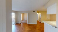 Located at 6425 Bankside Dr in Houston, TX, this single-floor residence offers a well-organized layout with a clean, neutral aesthetic throughout. The main living area is anchored by warm hardwood-style flooring that extends through the common spaces, creating a cohesive and inviting atmosphere. A large sliding glass door fitted with vertical blinds fills the living room with natural light and provides access to the outdoors. An open passage connects the living area to additional rooms, allowing for an easy flow through the home. A built-in shelving unit adds functional storage and a distinctive architectural detail to the living space. The galley-style kitchen is efficiently designed with ample white cabinetry lining both sides, a white appliance suite including a range and refrigerator, a countertop bar area that opens toward the living space, and hardwood-style flooring that ties the spaces together. The bedrooms feature neutral carpet, soft wall tones, and windows with blinds that allow for natural light and privacy. The bathroom is appointed with a wide vanity, generous counter space, white cabinetry, a well-lit mirror, and hardwood-style flooring. Throughout the home, consistent neutral wall tones and clean white trim create a bright, move-in-ready environment.