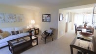 This two-story residence offers a thoughtfully arranged interior defined by a calm, neutral color palette throughout. The main living area serves as the heart of the home, featuring wall-to-wall carpeting in a warm beige tone that adds comfort underfoot. Soft cream-colored walls provide a clean backdrop, complemented by recessed ceiling details that lend a subtle architectural finish to the space.

The living area is well-proportioned, accommodating a seating arrangement that includes an accent chair with curved wooden arms and a glass-top coffee table, suggesting a balance of style and functionality. A framed wall piece adds a decorative focal point, while a table lamp introduces warm ambient lighting to the room.

An open corridor connects the living space to additional areas of the home, with a glimpse into what appears to be a brightly lit kitchen or utility space beyond. The hallway benefits from natural light filtering in from the far end, enhancing the overall brightness of the interior.

Spanning two floors, this residence provides versatile living arrangements with room to accommodate a variety of lifestyle needs. The cohesive design language—neutral finishes, soft flooring, and an open flow—creates a welcoming and livable environment throughout.