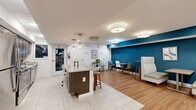 Located at 3600 Ely Pl SE in Washington, DC, this single-floor community space offers a well-appointed and inviting environment designed for gathering, relaxing, and socializing. The layout flows seamlessly across two primary zones — a kitchen/living area and a dedicated living room — creating a versatile and functional space throughout.

The kitchen is a standout feature, boasting rich dark wood cabinetry, a striking teal subway tile backsplash, stainless steel appliances including a refrigerator, gas range, and dishwasher, and a built-in wine rack. A large granite-topped island with wooden bar stools serves as a central hub for casual dining and conversation, illuminated by elegant pendant lighting.

The living and lounge areas are defined by a bold teal accent wall and warm wood-look flooring. A brick fireplace with a modern linear insert anchors one end of the living room, surrounded by colorful, contemporary lounge seating on a patterned area rug. Adjacent to this, a seating zone features high-back upholstered booth-style benches paired with compact tables — ideal for small group interactions or focused work.

Near the entry, a welcoming reception area with soft gray lounge chairs and large glass doors provides an open, accessible transition point. Flush-mounted drum lighting throughout ensures a bright, well-lit ambiance across all areas.