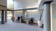 This sophisticated building lobby makes a striking first impression with its soaring double-height ceilings and expansive floor-to-ceiling windows that flood the space with natural light. The architectural design blends clean contemporary lines with warm, welcoming details, resulting in an ambiance that is both professional and inviting.

Upon entry, large glass double doors open into a spacious foyer anchored by a sleek, curved reception console with a frosted glass top surface, flanked by a pair of dark leather chairs. A framed mirror and decorative floral arrangements accent the console area, while oversized sculptural vases filled with tall decorative branches add organic texture and visual interest throughout.

The corridor extends deeper into the building, lined with structural columns and illuminated by a combination of recessed ambient lighting and subtle cove lighting that runs along accent walls. To one side, a built-in bank of resident mailboxes offers a practical and neatly integrated amenity. A welcoming message is displayed prominently on the wall, reinforcing the sense of community.

Lush greenery is positioned along an upper-level ledge overlooking the lobby, adding a refreshing natural element to the design. The flooring transitions between tile and carpet, with a durable entry mat at the main entrance. Curated artwork adorns the walls, completing the polished, well-appointed aesthetic of this communal space.