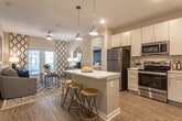 This well-designed single-floor residence offers a thoughtful open-concept layout that seamlessly connects the kitchen, dining, and living areas. The kitchen is a standout feature, equipped with stainless steel appliances including a refrigerator, range, microwave, and dishwasher. Crisp white cabinetry, a classic subway tile backsplash, and light quartz countertops complement the large center island with bar seating — ideal for casual dining or entertaining. Warm wood-look flooring and modern pendant lighting complete the kitchen's contemporary aesthetic. The living room is bright and inviting, benefiting from natural light through full-length windows and a glass door that opens to an outdoor area. A ceiling fan adds comfort to the space. The primary bedroom is generously sized with plush carpeting, large windows, and an en-suite bathroom featuring a quartz vanity, tiled walk-in shower, and direct access to a spacious walk-in closet with built-in shelving. A second bedroom provides additional flexibility and is similarly appointed with carpeted floors and natural light. A dedicated in-unit laundry room houses a full-size washer and dryer with overhead shelving for storage. Throughout, the neutral color palette and modern finishes create a cohesive, contemporary living environment.