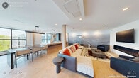 This contemporary single-floor space offers a thoughtfully designed open-plan layout that seamlessly integrates the living, dining, and kitchen areas into one cohesive environment. The entrance leads into a bright, airy interior finished with light hardwood flooring throughout, recessed lighting, and clean white ceilings that enhance the sense of openness.

The living area is anchored by a sleek linear fireplace set beneath a wall-mounted display, accompanied by a striking floor-to-ceiling decorative log storage feature that adds a natural, sculptural accent. A full-wall artistic mural in soft grey watercolor tones creates a dramatic backdrop behind the seating arrangement, which includes a plush sofa, accent chair, and a patterned area rug.

The kitchen is outfitted with flat-front powder-blue cabinetry, light stone countertops, a deep stainless steel sink, and integrated appliances including a built-in microwave and refrigerator drawers. A generous island with pendant lighting above serves as a natural divider between the kitchen and dining zone.

The entrance area features expansive floor-to-ceiling windows with black framing and a large sliding glass door that opens to an outdoor terrace, flooding the space with natural light and offering views of the surrounding exterior. A decorative river rock inset lines the base of the window wall, adding an organic design element to the otherwise modern aesthetic.