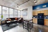 This single-floor loft-style space showcases a distinctive industrial aesthetic defined by exposed concrete ceilings, polished concrete floors, and an open-concept layout that seamlessly connects the main living areas. The living room is anchored by a large sectional sofa and benefits from an expansive wall of floor-to-ceiling windows fitted with horizontal blinds, flooding the space with abundant natural light and views of the surrounding urban environment. A ceiling fan with integrated lighting and exposed ductwork add to the raw, industrial character of the space.

The kitchen is fully equipped and well-appointed, featuring light maple cabinetry, dark countertops, a tile backsplash, and a full suite of black appliances including a range, microwave, dishwasher, and refrigerator. A bar-height dining table with stools serves as both a dining and casual workspace area, positioned at the boundary between the kitchen and living room.

The bedroom continues the industrial design language with a concrete accent wall serving as a textural backdrop. The room accommodates a full-sized bed with coordinating furniture and receives natural light through a horizontal clerestory window. A geometric metal shelving unit in the hallway adds a decorative and functional transitional element between spaces.