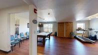 Located at 3475 American Dr in Colorado Springs, CO, this exercise and recreation room is a versatile, single-floor amenity space designed to serve both fitness and leisure needs. Rich hardwood flooring runs throughout, complementing the neutral-toned walls and textured ceilings that lend the room a warm, functional character.

The space is thoughtfully divided into two primary zones. The fitness area is equipped with a treadmill, stationary bike, elliptical trainer, and a rack of free weights, all positioned near a window that allows natural light to flow in. Track lighting overhead ensures the area remains well-lit for workouts at any time of day.

Transitioning into the recreation zone, a full-size pool table serves as the room's centerpiece, accompanied by a wall-mounted cue rack and a dartboard. A wall-mounted flat-screen TV and pendant lighting add to the social atmosphere of this area. A row of blue chairs lines one wall, providing ample seating for spectators or gatherings.

Storage doors along the perimeter offer practical utility, keeping the open floor plan clean and uncluttered. The combination of natural light, durable flooring, and dual-purpose layout makes this space a functional and inviting amenity for recreation and fitness alike.