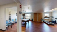Located at 3475 American Dr in Colorado Springs, CO, this exercise and recreation room is a versatile, single-floor amenity space designed to serve both fitness and leisure needs. Rich hardwood flooring runs throughout, complementing the neutral-toned walls and textured ceilings that lend the room a warm, functional character.

The space is thoughtfully divided into two primary zones. The fitness area is equipped with a treadmill, stationary bike, elliptical trainer, and a rack of free weights, all positioned near a window that allows natural light to flow in. Track lighting overhead ensures the area remains well-lit for workouts at any time of day.

Transitioning into the recreation zone, a full-size pool table serves as the room's centerpiece, accompanied by a wall-mounted cue rack and a dartboard. A wall-mounted flat-screen TV and pendant lighting add to the social atmosphere of this area. A row of blue chairs lines one wall, providing ample seating for spectators or gatherings.

Storage doors along the perimeter offer practical utility, keeping the open floor plan clean and uncluttered. The combination of natural light, durable flooring, and dual-purpose layout makes this space a functional and inviting amenity for recreation and fitness alike.