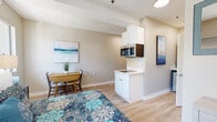 Located at 480 S El Camino Real in Encinitas, California, this single-floor studio is thoughtfully designed to balance comfort and functionality in a cohesive, open layout. Light wood-style flooring runs throughout, lending warmth and visual continuity to the entire space.

The sleeping area serves as the heart of the studio, accommodating a full-sized bed flanked by matching nightstands and bedside lamps. A tufted accent chair adds a touch of style, while multiple windows invite an abundance of natural light and offer pleasant views of the surrounding greenery. Coastal-inspired artwork adorns the walls, reinforcing a calm and serene ambiance throughout.

A compact kitchenette is seamlessly integrated into the living space, featuring white shaker-style cabinetry, an over-the-counter microwave, and a small cooktop surface — providing essential amenities without overwhelming the open floor plan. A drop-leaf dining table with chairs is positioned nearby, offering a dedicated spot for meals.

The bathroom is designed with accessibility in mind, featuring a walk-in shower equipped with grab bars, a built-in bench seat, and an emergency call panel. Light wood flooring continues here, maintaining design consistency.

Additional storage is provided via a dresser and closet area, ensuring practical organization within the studio's efficient footprint.