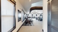 The Ovation at Town Madison Fitness Center at 300 Intergraph Way in Madison, Alabama, is a comprehensively equipped facility occupying a generously sized single floor. The space features soaring vaulted ceilings that lend an open, airy atmosphere, complemented by exposed industrial-style HVAC ductwork that adds a modern aesthetic contrast to the bright white walls and neutral-toned flooring throughout.

The facility is organized into distinct training zones. The cardio area lines one side of the main floor, housing a full row of treadmills, elliptical trainers, stationary cycles, and stair climbers, many positioned near large windows and glass doors that offer outdoor views and ample natural light. Wall-mounted televisions provide entertainment throughout this section.

A dedicated strength training zone features a comprehensive array of selectorized cable and resistance machines, accommodating a wide range of muscle groups. Adjacent to this is a functional training area equipped with power racks, barbells, weight plates, a full dumbbell rack, battle ropes, and a heavy punching bag, ideal for high-intensity and free-weight workouts.

One section of the facility includes glass panel garage-style doors that open to the outdoors, integrating the interior with exterior space. Ceiling fans and recessed lighting ensure comfort and visibility throughout. The rubberized flooring provides durability and safety across all training zones.