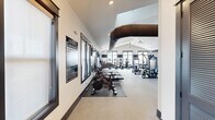 The Ovation at Town Madison Fitness Center at 300 Intergraph Way in Madison, Alabama, is a comprehensively equipped facility occupying a generously sized single floor. The space features soaring vaulted ceilings that lend an open, airy atmosphere, complemented by exposed industrial-style HVAC ductwork that adds a modern aesthetic contrast to the bright white walls and neutral-toned flooring throughout.

The facility is organized into distinct training zones. The cardio area lines one side of the main floor, housing a full row of treadmills, elliptical trainers, stationary cycles, and stair climbers, many positioned near large windows and glass doors that offer outdoor views and ample natural light. Wall-mounted televisions provide entertainment throughout this section.

A dedicated strength training zone features a comprehensive array of selectorized cable and resistance machines, accommodating a wide range of muscle groups. Adjacent to this is a functional training area equipped with power racks, barbells, weight plates, a full dumbbell rack, battle ropes, and a heavy punching bag, ideal for high-intensity and free-weight workouts.

One section of the facility includes glass panel garage-style doors that open to the outdoors, integrating the interior with exterior space. Ceiling fans and recessed lighting ensure comfort and visibility throughout. The rubberized flooring provides durability and safety across all training zones.