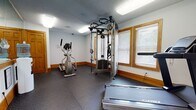Located at 9724 Virginia Centerway Pl in Glen Allen, Virginia, this single-floor amenity space houses a well-equipped fitness center and a laundry facility designed to serve its community. The fitness center features durable rubber flooring throughout, providing a safe and supportive surface for exercise. The room is outfitted with a selection of quality machines, including a Cybex treadmill with intelligent suspension technology, an elliptical cross-trainer, and a multi-function cable resistance machine with a full weight stack and suspension training attachments. Natural light filters in through wood-framed windows fitted with blinds, complementing the bright overhead fluorescent lighting. White walls with warm wood baseboards give the space a clean, functional aesthetic. A wall-mounted television and posted exercise guides add practical touches for users. A large mirror along one wall enhances the sense of space. Entry is through a classic wood-paneled door labeled "Fitness Center," with glass panel access to the exterior. Adjacent to the fitness area, the laundry room features light tile flooring, a front-load washer/dryer unit, a folding table, and an electrical panel, providing residents with essential on-site laundry convenience.