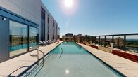 This space offers a compelling combination of outdoor luxury and functional interior amenities. The centerpiece is a generously sized rooftop lap pool, elegantly designed and framed by a spacious stone-tiled deck. Rows of cushioned lounge chairs line the poolside, providing ample space for relaxation under open skies. The rooftop setting delivers sweeping panoramic city views in all directions, enclosed by low-profile glass railings that preserve the unobstructed sightlines. The surrounding modern building architecture, featuring large windows and clean geometric lines, lends a polished, contemporary character to the outdoor environment.

Inside, the space includes a half bathroom finished in a classic, understated style. The vanity features a wood cabinet base with a light countertop and a framed mirror above, accented by warm brass hardware and fixtures throughout, including switch plates and outlet covers that lend a cohesive traditional touch. An over-toilet shelving unit provides additional storage, keeping the compact space organized and functional. Neutral wall tones and simple, clean surfaces contribute to a calm, no-fuss aesthetic. Together, the rooftop amenity and interior bathroom create a well-rounded space that balances outdoor leisure with practical indoor convenience.