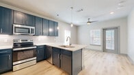 This single-floor space presents a cohesive, contemporary design characterized by a neutral color palette accented with striking navy blue cabinetry throughout. Light wood-look flooring runs continuously through all areas of the unit, creating a seamless and unified aesthetic.

The kitchen serves as a central feature of the space, showcasing bold navy shaker-style cabinetry that extends to the upper walls, providing generous storage. A peninsula with a white quartz countertop extends into the living area, fitted with an undermount sink and brushed nickel faucet. Stainless steel appliances, including a refrigerator with ice and water dispenser, electric range, microwave, and dishwasher, complement the modern finishes. Decorative pendant lights hang above the peninsula, while a white subway tile backsplash adds texture to the kitchen walls.

The living and entry areas are bright and open, benefiting from multiple windows fitted with white horizontal blinds that invite natural light. A ceiling fan circulates air throughout the main living space. A glass-panel entry door further enhances natural light in the foyer area.

The bathroom continues the navy cabinetry theme, featuring a matching vanity with a white countertop, an overhead storage cabinet, a framed mirror, and a shower-tub combination. Grab bars and practical hardware complete the well-appointed bathroom.