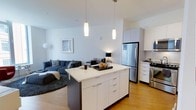 This single-floor unit offers a well-designed open-concept layout that seamlessly connects the kitchen and living areas. The kitchen serves as a central focal point, anchored by a large island with a crisp white countertop, ample drawer storage, and a dark base that provides a refined contrast. Pendant lighting hangs above the island, adding both task illumination and a modern aesthetic touch. The kitchen is equipped with stainless steel appliances, including a full-size refrigerator and range, complemented by white cabinetry that extends to the upper walls for generous storage. Light hardwood flooring runs throughout, contributing to a warm, cohesive feel. The adjoining living area provides comfortable space for relaxing, featuring neutral-toned walls that enhance the sense of openness and brightness. High ceilings amplify the airy atmosphere throughout the unit. The overall design aesthetic is clean and contemporary, with a neutral color palette, modern fixtures, and an efficient use of space that makes this one-bedroom, one-bathroom unit both functional and visually appealing.