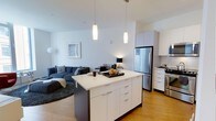 This single-floor unit offers a well-designed open-concept layout that seamlessly connects the kitchen and living areas. The kitchen serves as a central focal point, anchored by a large island with a crisp white countertop, ample drawer storage, and a dark base that provides a refined contrast. Pendant lighting hangs above the island, adding both task illumination and a modern aesthetic touch. The kitchen is equipped with stainless steel appliances, including a full-size refrigerator and range, complemented by white cabinetry that extends to the upper walls for generous storage. Light hardwood flooring runs throughout, contributing to a warm, cohesive feel. The adjoining living area provides comfortable space for relaxing, featuring neutral-toned walls that enhance the sense of openness and brightness. High ceilings amplify the airy atmosphere throughout the unit. The overall design aesthetic is clean and contemporary, with a neutral color palette, modern fixtures, and an efficient use of space that makes this one-bedroom, one-bathroom unit both functional and visually appealing.