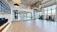 Located at 2130 Arapahoe St in Denver's vibrant RiNo neighborhood, this contemporary fitness studio offers a thoughtfully designed, single-floor layout optimized for group exercise, yoga, and strength training. The space is defined by its industrial-modern aesthetic, featuring exposed concrete columns, visible ductwork and piping overhead, and soaring ceilings that create an expansive, open atmosphere throughout.

Large, floor-to-ceiling windows line the perimeter, flooding the studio with abundant natural light and offering views of the surrounding Denver cityscape. Light hardwood flooring runs throughout, complementing the bright, airy ambiance and providing a functional surface for a variety of fitness activities.

The main studio area is generously proportioned, accommodating rows of yoga mats and offering ample space for movement-based classes. Full-length mirrors are strategically positioned along the walls to support proper form during workouts. A dedicated strength training zone includes a well-stocked dumbbell rack, while built-in shelving near the entrance provides organized storage for rolled yoga mats and equipment.

Linear pendant lighting and track fixtures are suspended from the exposed ceiling, delivering consistent, well-distributed illumination across all areas. Roller shades on the windows allow for light control during classes. The overall design balances raw industrial character with clean, modern finishes to create a motivating and professional environment.