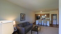 This single-floor residence offers a thoughtfully designed open-concept layout that integrates the living and kitchen areas into one cohesive, functional space. The living area is warm and inviting, featuring plush carpeting, neutral-toned walls, and ample room for comfortable seating. A framed piece of wall art adds a decorative touch, while a floor lamp contributes to the cozy ambiance.

The space flows naturally into the kitchen, which is equipped with warm wood-toned cabinetry, black appliances including a refrigerator and range, and a light-colored countertop that provides generous prep space. A peninsula-style breakfast bar separates the kitchen from the living area, furnished with tall bar-height chairs — ideal for casual dining or entertaining. Pendant lights hang above the kitchen area, offering both task lighting and a stylish design element.

The open floor plan creates a sense of spaciousness, with clean sightlines throughout. The combination of carpet in the living zone and hard flooring in the kitchen defines each area while maintaining a unified aesthetic. Overall, the space presents a comfortable, practical, and well-lit environment suited for everyday living.