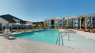 Located at 8251 Windsor Hill Blvd in North Charleston, SC, this outdoor amenity space is designed to deliver a resort-style experience within a well-appointed residential community. At its centerpiece sits a generously sized swimming pool with crystal-clear water, framed by a broad concrete sundeck outfitted with multiple chaise lounges and shaded umbrella stations — ideal for relaxing under the South Carolina sun.

A covered patio area extends along one side of the pool, featuring large floor-to-ceiling glass windows and wide glass doors that blur the boundary between indoor and outdoor space. This sheltered zone is furnished with wicker lounge chairs and wooden dining sets, offering comfortable seating for outdoor gatherings in any weather. The patio's white-painted columns and board-and-batten siding lend a coastal cottage aesthetic that complements the surrounding multi-story residential buildings, which are clad in a classic blue-and-white palette.

Lush tropical plantings, including palm trees and ornamental grasses, are thoughtfully integrated throughout the space, adding greenery and a sense of privacy. The overall design balances modern outdoor living with a relaxed coastal charm, creating an inviting communal environment well-suited to the warm climate of the South Carolina Lowcountry.