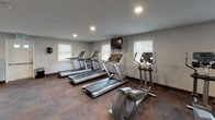 This fitness center is a thoughtfully arranged, single-floor space designed to accommodate a range of workout needs. The main exercise area is generously sized, featuring patterned carpet tile flooring throughout and recessed lighting in smooth, neutral-toned ceilings. Multiple windows fitted with horizontal blinds allow natural light to filter in, creating a bright and functional environment.

The cardio section is well-stocked with several treadmills and elliptical trainers positioned along the walls, complemented by a wall-mounted flat-screen television for entertainment during workouts. A dedicated strength training zone includes a multi-station cable machine, a weight bench, and a rack of kettlebells, offering versatile resistance training options. A stationary bike and a water cooler are also present, adding to the practicality of the space.

One wall features a bold navy blue accent, adding visual contrast to the otherwise neutral grey palette. A white open-cube shelving unit sits against this accent wall, providing organizational storage. A Pepsi vending machine is conveniently positioned nearby. The space is accessible via an interior door and is designed to serve as a shared-use fitness facility.