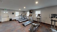 This fitness center is a thoughtfully arranged, single-floor space designed to accommodate a range of workout needs. The main exercise area is generously sized, featuring patterned carpet tile flooring throughout and recessed lighting in smooth, neutral-toned ceilings. Multiple windows fitted with horizontal blinds allow natural light to filter in, creating a bright and functional environment.

The cardio section is well-stocked with several treadmills and elliptical trainers positioned along the walls, complemented by a wall-mounted flat-screen television for entertainment during workouts. A dedicated strength training zone includes a multi-station cable machine, a weight bench, and a rack of kettlebells, offering versatile resistance training options. A stationary bike and a water cooler are also present, adding to the practicality of the space.

One wall features a bold navy blue accent, adding visual contrast to the otherwise neutral grey palette. A white open-cube shelving unit sits against this accent wall, providing organizational storage. A Pepsi vending machine is conveniently positioned nearby. The space is accessible via an interior door and is designed to serve as a shared-use fitness facility.