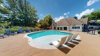 Located at Highland Lake Circle in Decatur, Georgia, this outdoor amenity area is a well-appointed residential retreat designed for relaxation and leisure. At the heart of the space is a generously sized freeform swimming pool with crystal-clear turquoise water, gentle entry steps, and built-in in-pool lounge chairs that invite guests to unwind at the water's edge.

The surrounding sundeck is finished in warm-toned concrete pavers and lined with rows of sleek chaise lounges, providing ample space for sunbathing. A shaded seating area with a market umbrella and outdoor dining furniture offers a comfortable alternative for those seeking respite from the sun.

A charming stone-accented clubhouse anchors one end of the pool area, featuring a prominent stone chimney, a pitched shingle roof, and large French doors that open directly onto the patio — seamlessly connecting indoor and outdoor spaces. On the opposite end of the pool deck, a handsome wooden pergola serves as a focal point, framing an additional lounge and dining area perfect for gatherings.

The space is richly landscaped with mature oak trees, flowering crepe myrtles in full bloom, ornamental grasses, and layered shrubs that create a lush, park-like atmosphere. The natural greenery provides both privacy and shade, giving the entire area a serene, resort-like ambiance.