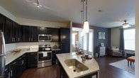This single-floor residence showcases a thoughtfully designed open-concept layout that seamlessly connects the kitchen and living areas, creating a spacious and cohesive environment. The kitchen is a standout feature, outfitted with dark espresso cabinetry that extends from floor to ceiling, offering generous storage throughout. Granite countertops with a speckled finish add a refined touch, complemented by a large undermount stainless steel sink set within a central island. Stainless steel appliances, including an electric range and built-in microwave, lend a sleek, contemporary feel. Elegant pendant lighting hangs above the island, providing both task illumination and a striking visual accent. Track lighting along the ceiling further enhances the kitchen's functionality. The dark hardwood flooring flows consistently throughout, anchoring the space with warmth and continuity. Adjacent to the kitchen, the living area benefits from an abundance of natural light streaming through large windows fitted with horizontal blinds. Accent walls in deep slate blue add depth and character to the interior. The overall ambiance is modern and polished, with a careful balance of dark and light tones that creates an inviting yet sophisticated atmosphere throughout the home.