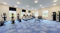 Located at 24 Bay Front Dr in Traverse City, Michigan, this single-floor fitness center is a well-appointed facility designed to support a comprehensive workout experience. The space is divided into distinct exercise zones, each thoughtfully outfitted and finished to promote both function and comfort.

The main cardio and strength area features a geometric-patterned blue carpet floor, creating a visually dynamic backdrop for an array of treadmills, elliptical trainers, and cable machines. A full wall of mirrors amplifies the sense of space while allowing users to monitor their form, and multiple wall-mounted flat-screen televisions are positioned throughout for entertainment during workouts. Large windows invite natural light and provide views of the surrounding greenery.

A dedicated free weight section includes a dumbbell rack and adjustable bench, while a separate area accommodates stability balls, kettlebells, and suspension training equipment neatly organized on racks. Built-in shelving along one wall offers convenient storage space.

A secondary room, finished with light neutral carpet, serves as a dedicated cycling and rowing studio. This bright, windowed space houses stationary bikes and a rowing machine, offering a quieter, focused environment for low-impact cardio training.

The entry vestibule features glass double doors, smooth transitions between zones, and clean, modern finishes throughout, making this fitness center a functional and welcoming space for users of all fitness levels.
