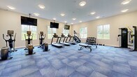 Located at 24 Bay Front Dr in Traverse City, Michigan, this single-floor fitness center is a well-appointed facility designed to support a comprehensive workout experience. The space is divided into distinct exercise zones, each thoughtfully outfitted and finished to promote both function and comfort.

The main cardio and strength area features a geometric-patterned blue carpet floor, creating a visually dynamic backdrop for an array of treadmills, elliptical trainers, and cable machines. A full wall of mirrors amplifies the sense of space while allowing users to monitor their form, and multiple wall-mounted flat-screen televisions are positioned throughout for entertainment during workouts. Large windows invite natural light and provide views of the surrounding greenery.

A dedicated free weight section includes a dumbbell rack and adjustable bench, while a separate area accommodates stability balls, kettlebells, and suspension training equipment neatly organized on racks. Built-in shelving along one wall offers convenient storage space.

A secondary room, finished with light neutral carpet, serves as a dedicated cycling and rowing studio. This bright, windowed space houses stationary bikes and a rowing machine, offering a quieter, focused environment for low-impact cardio training.

The entry vestibule features glass double doors, smooth transitions between zones, and clean, modern finishes throughout, making this fitness center a functional and welcoming space for users of all fitness levels.