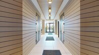 This thoughtfully designed amenity floor opens with a sleek entry corridor finished in light horizontal-slat wall paneling, large-format white tile flooring, and a series of gold pendant light fixtures that draw the eye toward glass double doors opening to the outdoors. Secure keypad access points are integrated seamlessly into the corridor design.

The resident lounge is a bright, welcoming gathering space featuring wide-plank hardwood flooring, recessed cove lighting, and floor-to-ceiling glass walls with French doors that open directly onto a landscaped outdoor courtyard. The lounge is divided into distinct zones, including a comfortable seating area with a large wall-mounted display and built-in shelving, as well as a casual dining and co-working area set beneath a statement pendant fixture with soft blue botanical wallcovering as an accent.

The fitness center is a standout feature, generously proportioned and outfitted with a comprehensive range of strength and cardio equipment. Rubber flooring, linear pendant lighting, textured wall finishes, and floor-to-ceiling mirrors create a professional, high-end gym atmosphere. An adjoining flex studio space, lined with mirrored walls and its own mounted display, provides room for stretching, yoga, or group training. Together, these amenities deliver a cohesive, upscale experience throughout.