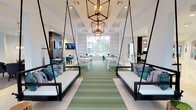 This beautifully designed single-floor amenity lounge offers a one-of-a-kind experience that blends comfort, style, and functionality. The space is immediately defined by its most striking feature: suspended swing sofas anchored from the ceiling by bold black metal frames, offering a playful yet sophisticated seating experience. Plush white cushions and teal and navy accent pillows complement the room's coastal-inspired color palette. Overhead, a series of geometric lantern-style pendant light fixtures hang from a refined coffered ceiling, adding warmth and architectural interest. A striped green area rug runs the length of the central corridor, grounding the space and guiding the eye toward the far end of the room, where large windows flood the interior with natural light and offer views of lush greenery outside. The background wall features a bold, patterned wallcovering that adds texture and visual depth. Beyond the central lounge area, dining tables with teal chairs provide a functional workspace or gathering spot, while a wall-mounted television and additional seating areas extend the space's versatility. The overall ambiance is bright, airy, and welcoming — a well-appointed retreat designed for relaxation and social connection.