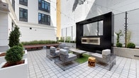 This outdoor courtyard showcases a thoughtfully designed urban retreat nestled between multi-story buildings. The space is anchored by a bold matte-black structure housing a contemporary linear fireplace with a stainless steel insert, providing both a visual focal point and a functional heat source for cooler evenings. Overhead, strands of string lights are draped across the courtyard, adding a warm, ambient glow suited for evening gatherings.

Seating is arranged in conversational groupings, featuring modern wire-frame lounge chairs and sofas with light gray upholstered cushions, complemented by a low concrete coffee table. Rustic wood stump side tables add an organic contrast to the industrial wire aesthetic. A patch of artificial turf beneath the seating area introduces a touch of greenery, while raised planters along the perimeter contain ornamental grasses and trimmed shrubs, softening the hardscape.

The tiled ground surface is laid in a clean grid pattern, reinforcing the space's structured, contemporary feel. A large-scale bird mural adorns one of the surrounding building walls, serving as a dramatic artistic backdrop. Wooden benches along the perimeter offer additional informal seating, completing this well-appointed communal outdoor space.