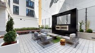 This outdoor courtyard showcases a thoughtfully designed urban retreat nestled between multi-story buildings. The space is anchored by a bold matte-black structure housing a contemporary linear fireplace with a stainless steel insert, providing both a visual focal point and a functional heat source for cooler evenings. Overhead, strands of string lights are draped across the courtyard, adding a warm, ambient glow suited for evening gatherings.

Seating is arranged in conversational groupings, featuring modern wire-frame lounge chairs and sofas with light gray upholstered cushions, complemented by a low concrete coffee table. Rustic wood stump side tables add an organic contrast to the industrial wire aesthetic. A patch of artificial turf beneath the seating area introduces a touch of greenery, while raised planters along the perimeter contain ornamental grasses and trimmed shrubs, softening the hardscape.

The tiled ground surface is laid in a clean grid pattern, reinforcing the space's structured, contemporary feel. A large-scale bird mural adorns one of the surrounding building walls, serving as a dramatic artistic backdrop. Wooden benches along the perimeter offer additional informal seating, completing this well-appointed communal outdoor space.