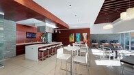 This single-floor space showcases a contemporary design defined by a striking contrast between rich, dark wood paneling and crisp white surfaces. The open layout integrates several functional zones seamlessly. A fully equipped kitchen anchors one side of the space, featuring stainless steel appliances, under-cabinet range hood, and a long white countertop island lined with modern black-and-white bar stools. Pendant lighting in polished chrome hangs above the kitchen area, adding a refined touch.

Adjacent to the kitchen, multiple high-top bistro tables with white chairs are arranged to encourage social interaction, bathed in natural light streaming through large windows. The light tile flooring throughout enhances the bright, airy atmosphere and complements the bold wood elements overhead and on the walls.

A workstation area is visible toward the back, equipped with computers and monitors, suggesting a business or co-working function. Colorful contemporary artwork adorns the wood-paneled walls, adding vibrancy and character. The angular ceiling design, incorporating both white drywall and warm wood soffits, adds architectural interest and depth to the overall ambiance.