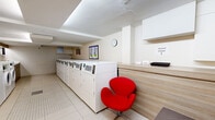 Located at 3470 Rue Stanley in downtown Montréal, this well-appointed shared laundry facility offers a clean, functional, and thoughtfully designed space for residents. The room is laid out efficiently, with rows of commercial-grade front-load washers and top-load dryers lining both sides, providing ample laundry capacity. A dedicated utility area features a built-in stainless steel sink set into warm wood-toned cabinetry with a white countertop, complemented by a large mirror above — ideal for hand-washing or pre-treating garments. Digital information displays are mounted on the walls, offering users convenient access to machine instructions or service details. The flooring throughout consists of light-toned ceramic tiles, contributing to the bright and hygienic atmosphere. Recessed ceiling panels provide even, diffused lighting across the entire space. A comfortable waiting area is furnished with distinctive red accent chairs and a small bench, offering a welcoming spot to relax. A corkboard and framed artwork add a personable touch to the otherwise utilitarian setting. The space is also equipped with a fire extinguisher and features a glazed door entrance, ensuring both safety and accessibility.