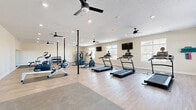 Located at 3555 Sirocco Way in Lafayette, Indiana, this well-appointed fitness center offers a generous, open single-floor layout designed to accommodate a wide variety of workout routines. The space features durable wood-style flooring throughout the main exercise area, complemented by rubberized mat sections that provide dedicated zones for cardio and strength training equipment.

The cardio section is equipped with multiple treadmills, elliptical machines, stationary bikes, and a rowing machine, providing ample options for aerobic exercise. A multi-functional cable machine and a rack of kettlebells and free weights, along with stability balls, round out the strength and functional training offerings. Wall-mounted flat-screen TVs are positioned for easy viewing from the cardio equipment, enhancing the workout experience.

Natural light streams in through several large windows along the walls, creating a bright and energizing atmosphere. Multiple ceiling fans are distributed throughout the space, ensuring consistent airflow and comfort. A full-length mirror on one wall allows users to monitor their form during exercise. Recessed lighting provides additional illumination throughout the room. A bulletin board near the entrance serves as an informational hub for users. The clean, neutral-toned walls and open floor plan contribute to an inviting and functional environment suited for daily fitness activity.