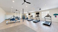 Located at 3555 Sirocco Way in Lafayette, Indiana, this well-appointed fitness center offers a generous, open single-floor layout designed to accommodate a wide variety of workout routines. The space features durable wood-style flooring throughout the main exercise area, complemented by rubberized mat sections that provide dedicated zones for cardio and strength training equipment.

The cardio section is equipped with multiple treadmills, elliptical machines, stationary bikes, and a rowing machine, providing ample options for aerobic exercise. A multi-functional cable machine and a rack of kettlebells and free weights, along with stability balls, round out the strength and functional training offerings. Wall-mounted flat-screen TVs are positioned for easy viewing from the cardio equipment, enhancing the workout experience.

Natural light streams in through several large windows along the walls, creating a bright and energizing atmosphere. Multiple ceiling fans are distributed throughout the space, ensuring consistent airflow and comfort. A full-length mirror on one wall allows users to monitor their form during exercise. Recessed lighting provides additional illumination throughout the room. A bulletin board near the entrance serves as an informational hub for users. The clean, neutral-toned walls and open floor plan contribute to an inviting and functional environment suited for daily fitness activity.